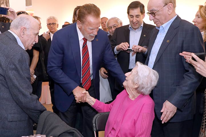 Arnold Schwarzenegger in a blue suit shakes the hand of a woman wearing a pink shirt