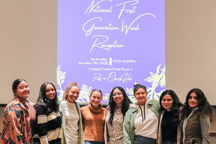 A group of 8 first-generation students in front of a screen announcing National First Generation Week