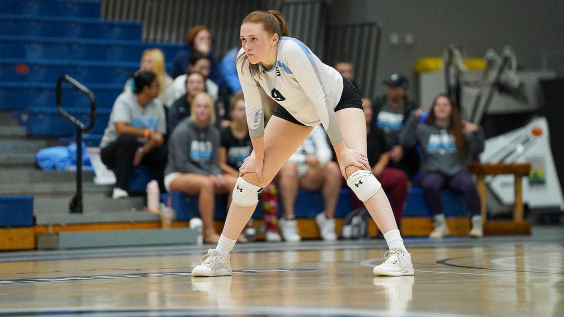 Kate Louer in white volleyball uniform stands with her hands on her knees on a volleyball court