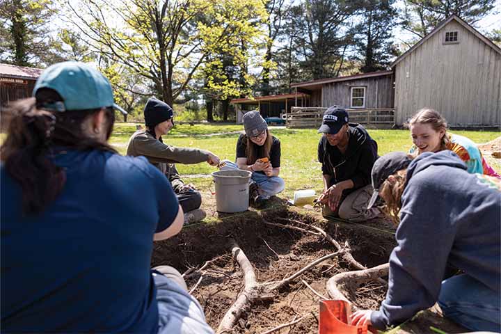 A group of students sitting on the ground with shovels and tools to examine a large set of tree roots