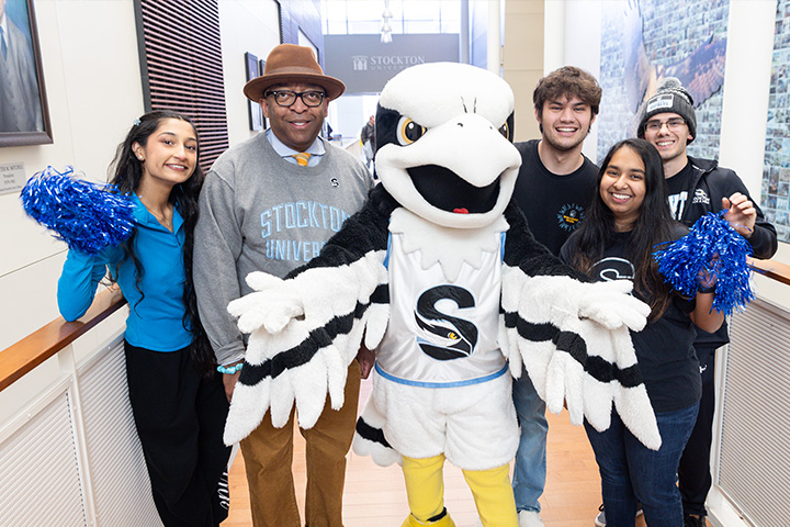 Staff and students pose with the Stockton Osprey mascot with pom poms on the bridge in the Campus Center