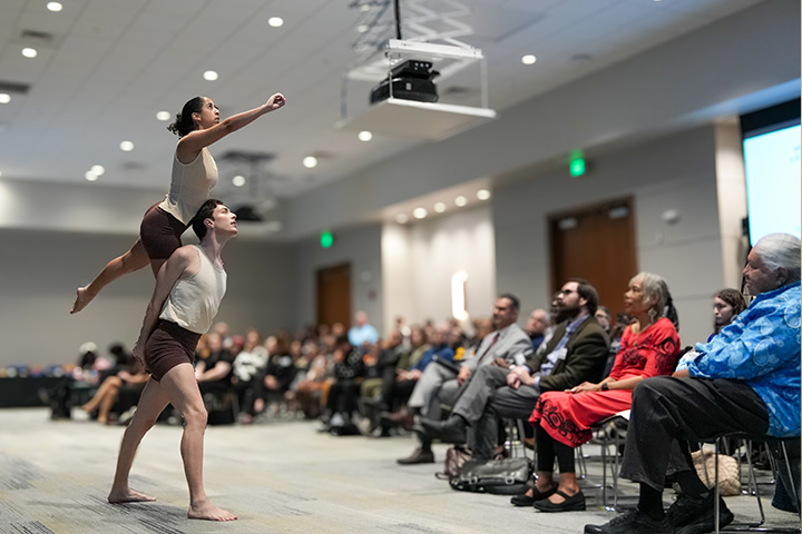 Two Stockton dance students in brown shorts and white shirts perform while a crowd watches in the Fannie Lou Hamer Event Room