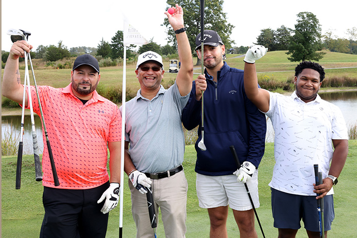 Four golfers stand on a golf course and hold up their clubs