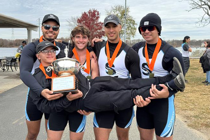 Four members of the men's rowing team hold up another team member horizontally who is holding a trophy