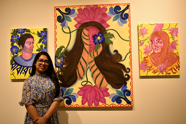A student in floral dress stands in front of her painted artwork hanging in the art gallery