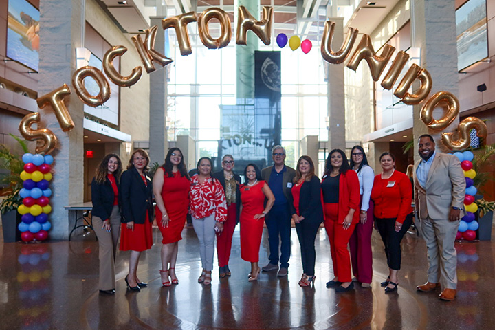 A group of Hispanic faculty and staff wear read and stand under a balloon arch spelling out Stockton Unidos