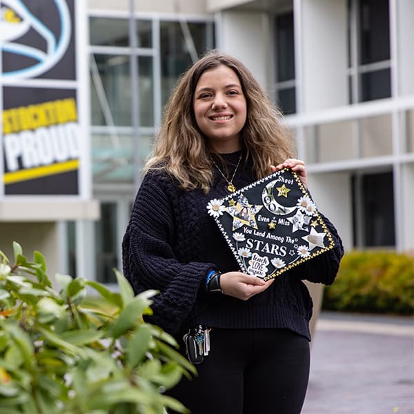 Angelina Maffei in front of the academic spine, holding her decorated graduation cap