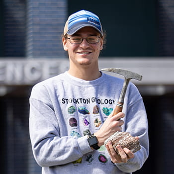 Joseph Rosal smiling and holding a rock and hammer in front of the Arts & Sciences building