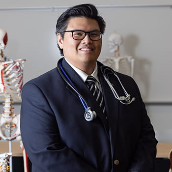 Matthew Romen smiling and sitting in a laboratory with a skeleton behind him