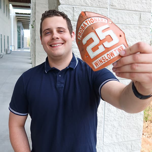 Ryan Bowie standing outside of the Campus Center, holding up a badge that says "Probationary, 25, Winslow Township"