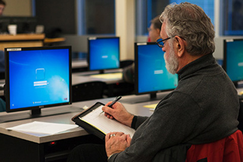 An older student at a computer, taking notes on pen and paper