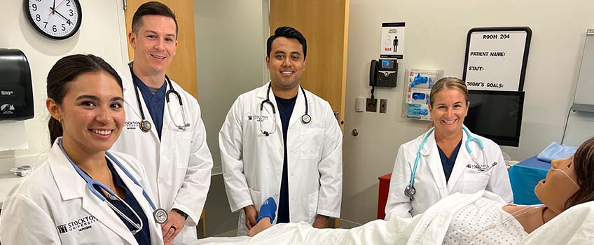 Four nursing students in white lab coats in a mock patient room