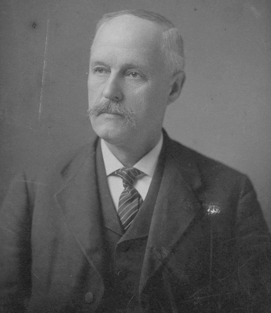 A grayscale photograph of a moustached man looking to the side of the photograph.