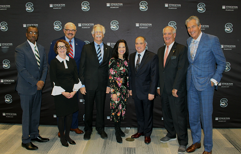 Panel, moderator, speaker and Mr. Levenson in front of University Logo Backdrop