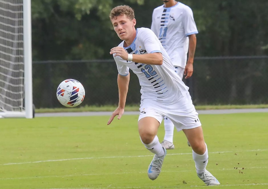Stockton soccer player in jersey chases ball on field during match, with teammate visible behind.