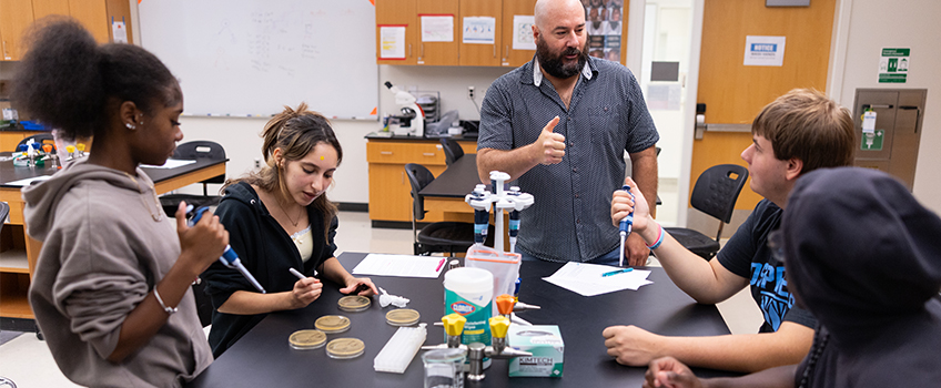 Students in a microbiology lab use pipettes and petri dishes while teacher gives instructions.