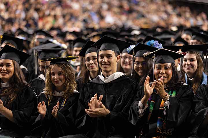 A group of graduates in black caps and gowns sit in Boardwalk Hall