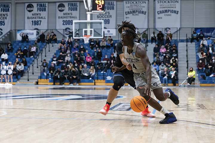 DJ Campbell wearing a gray Stockton jersey dribbling past a defender